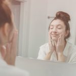 Young woman gently washing her face with cleanser while smiling and looking in a mirror