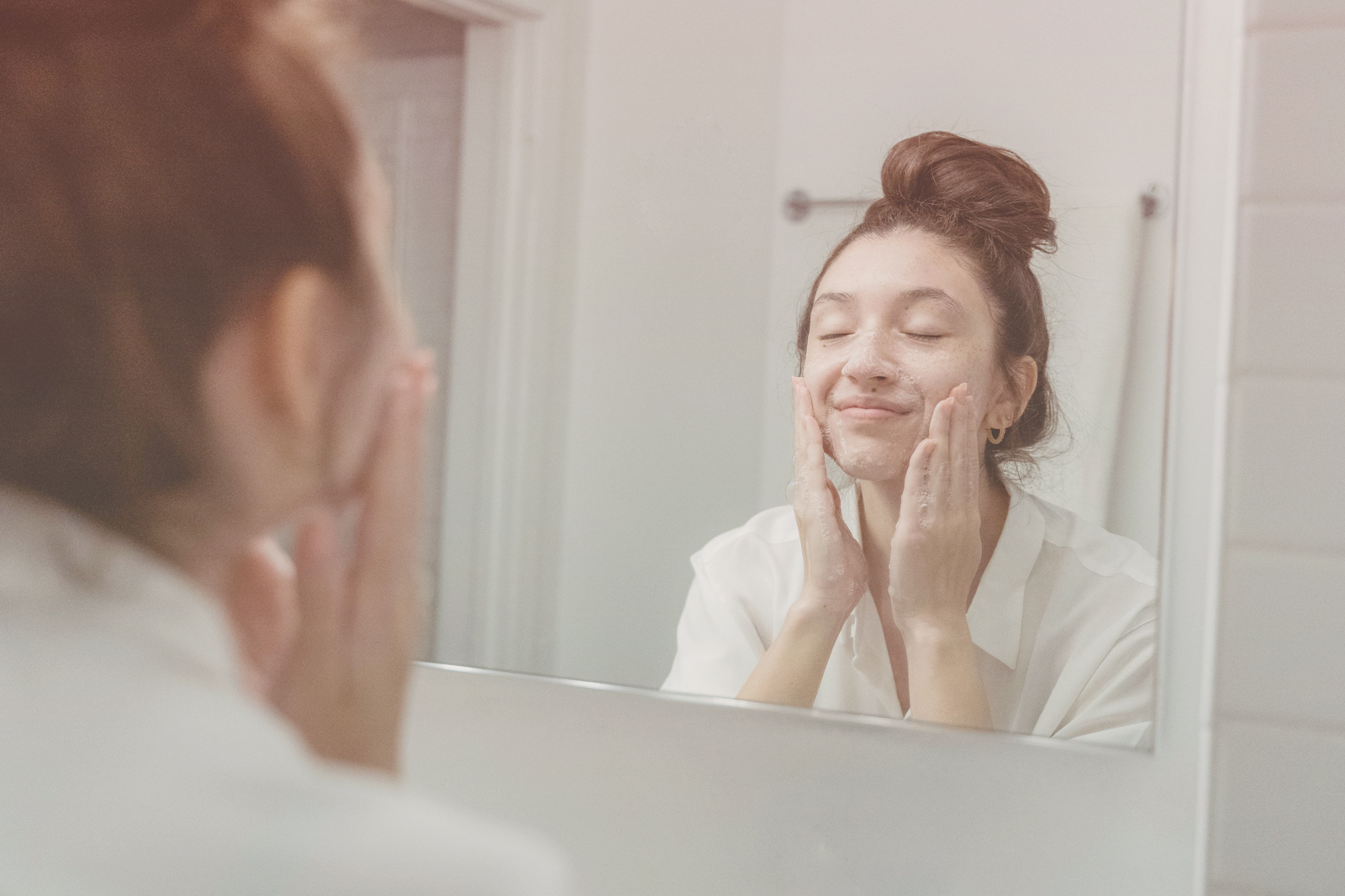 Young woman gently washing her face with cleanser while smiling and looking in a mirror