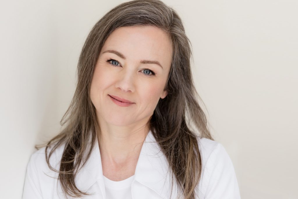 Sarah, RN, owner of a plasma aesthetics practice, smiling softly while leaning against a wall