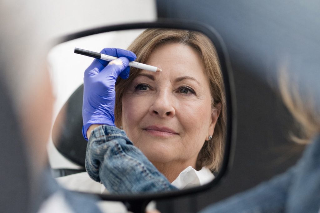 Middle aged woman viewing her skin in a mirror during a NeoGen consultation as practitioner performs facial assessment