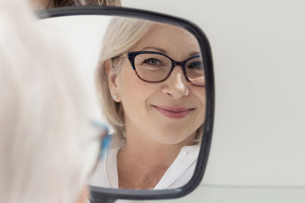 Midlife woman smiling while looking in a mirror, representing confidence and healthy skin