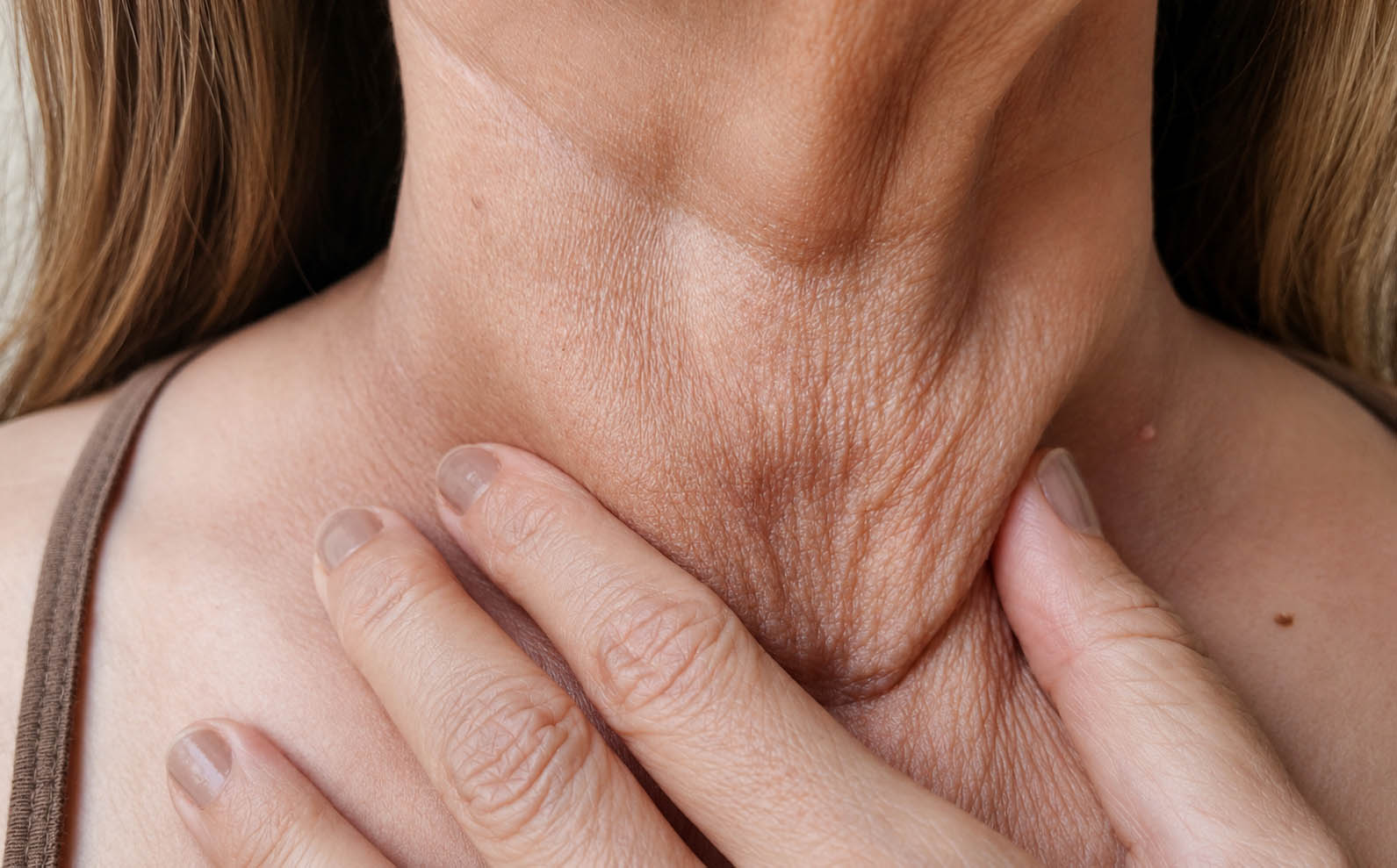 Close-up of a woman gently pinching loose, crepey skin on her neck, demonstrating neck laxity and the visible signs of collagen and elastin loss with aging.